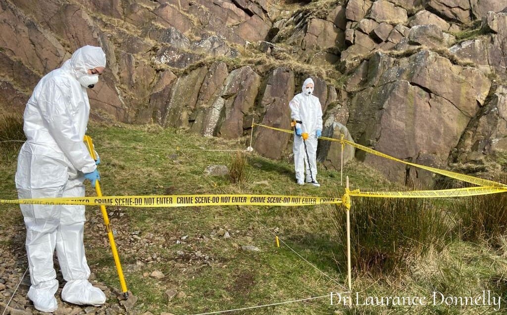 A person uses a metal detector or survey device on grassy moorland, with white marker tape laid across the ground and rocky hills behind them.