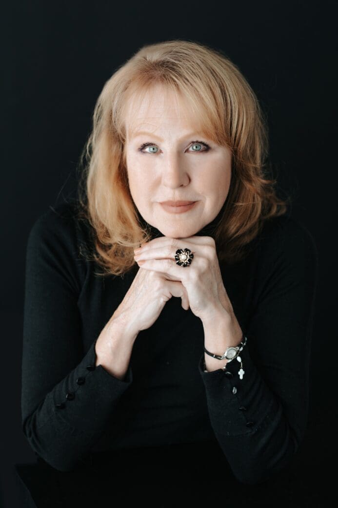 Studio portrait of Corinna Lain, wearing a black top, resting her chin on her hands against a dark background.
