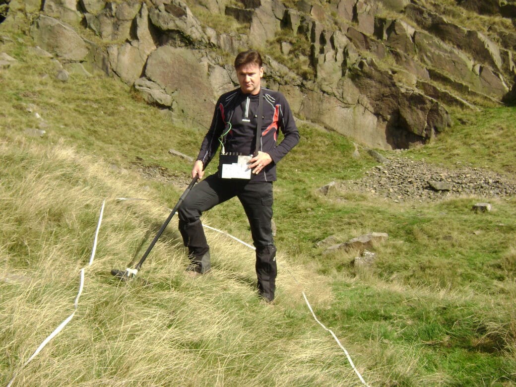 A person uses a metal detector or survey device on grassy moorland, with white marker tape laid across the ground and rocky hills behind them.