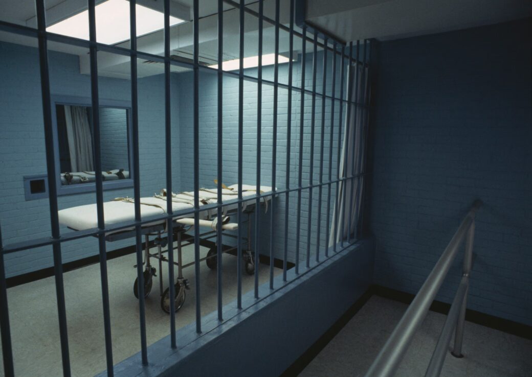 An empty execution chamber with a gurney behind metal bars, viewed from a dim corridor.