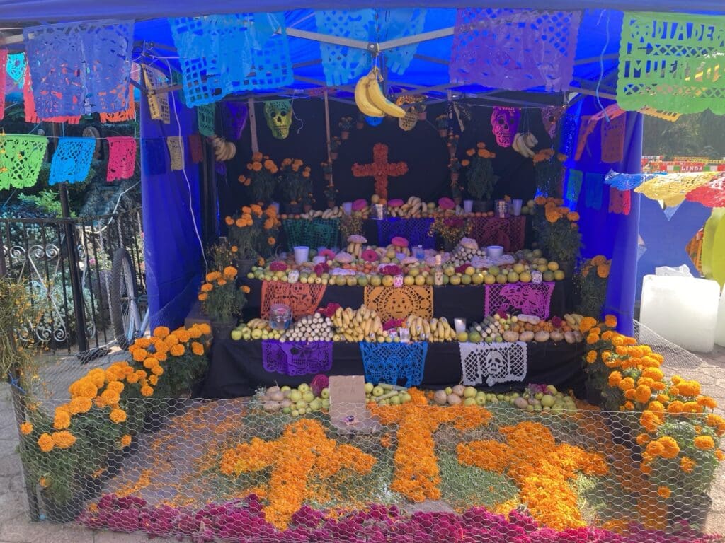 A Day of the Dead display. There are crosses made of orange flowers, papel picado paper cut outs across an altar, bananas hanging from a display above, food and skulls.