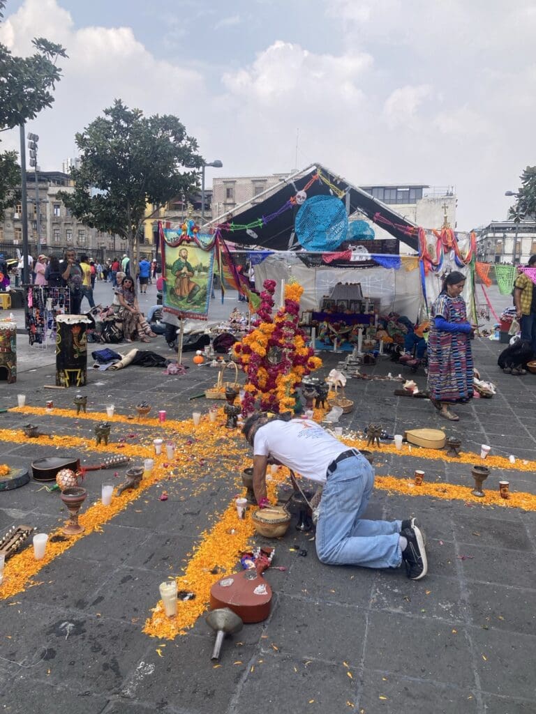 A Day of the Dead display. A man is crouching down. There are orange petals and instruments on the floor in the formation of an X (or plus sign.) There are many people behind the display.