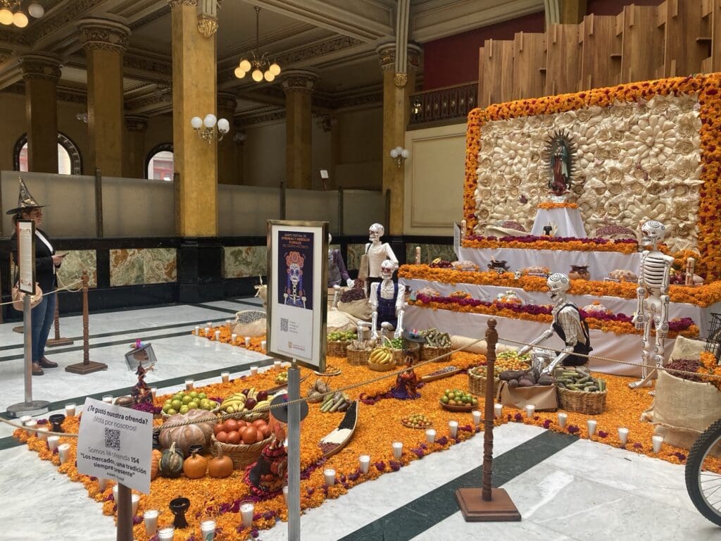 A Day of the Dead display. There are many orange petals on the ground, and food (such as fruits and vegetables) in bowls. There are also statues of skulls. This display is indoors.