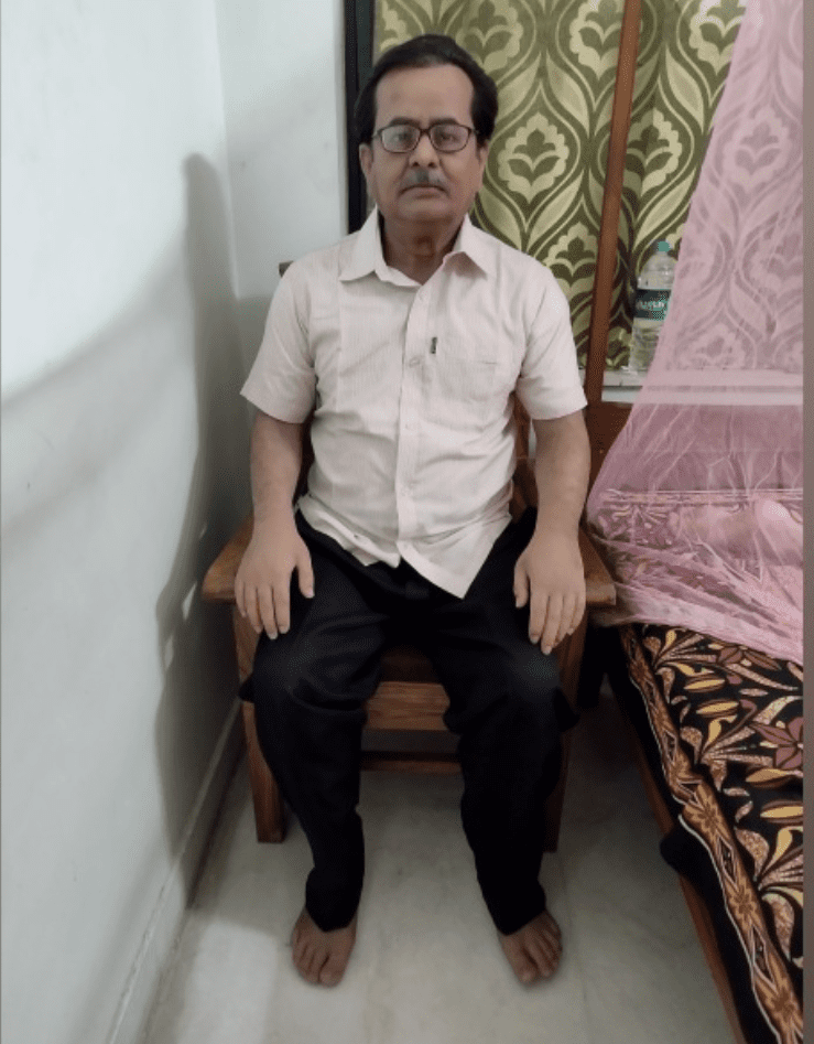 A life-size silicone memorial figure of a man seated on a wooden chair in a bedroom, wearing glasses, a pale shirt and dark trousers.