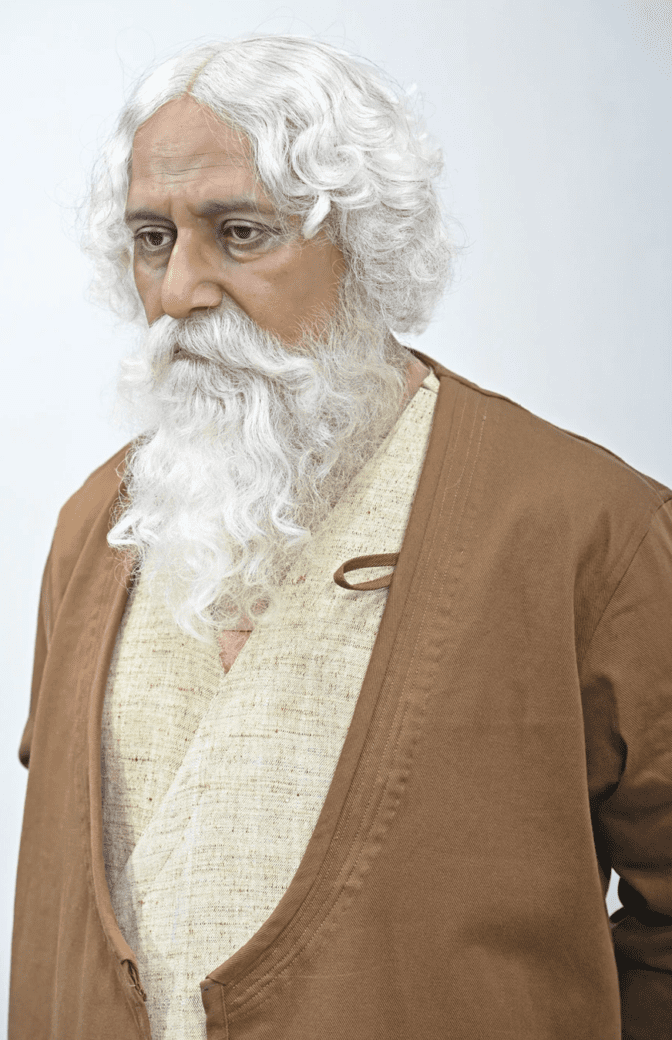 A hyper-realistic sculpture of an older man with white hair and a long white beard, wearing a cream outfit and brown outer robe against a plain background.