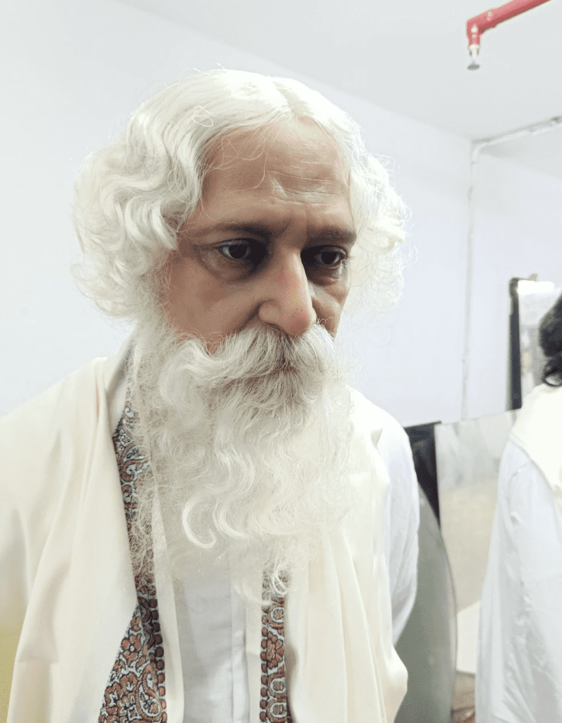 A hyper-realistic sculpture of an older man with white hair and a long white beard, wearing a cream outfit and brown outer robe against a plain background.