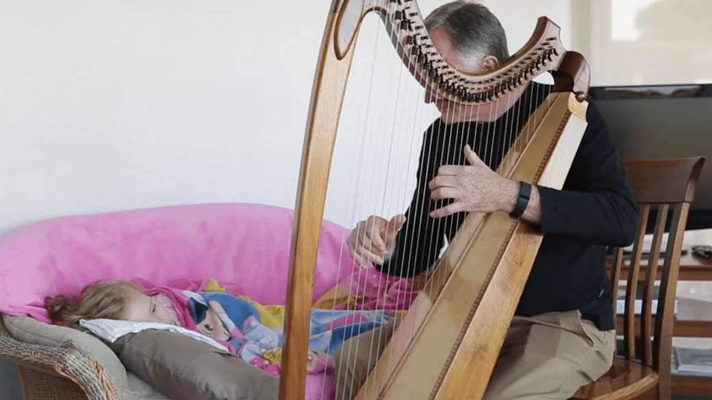 Peter Roberts sitting down on a wooden chair playing the Harp for a sick child. The child is wrapped in a pink blanket and is lying on a sofa next to the wooden chair.