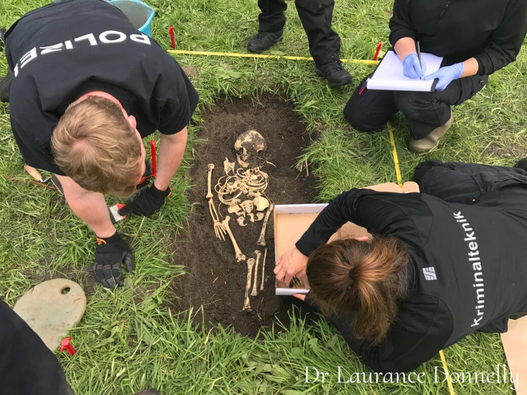 Forensic investigators carefully excavate skeletal remains from a marked area of grass, with one officer taking notes beside the scene.