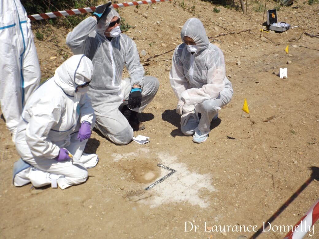 Forensic investigators in protective suits work behind police tape at an outdoor search scene, with evidence markers placed across the ground.