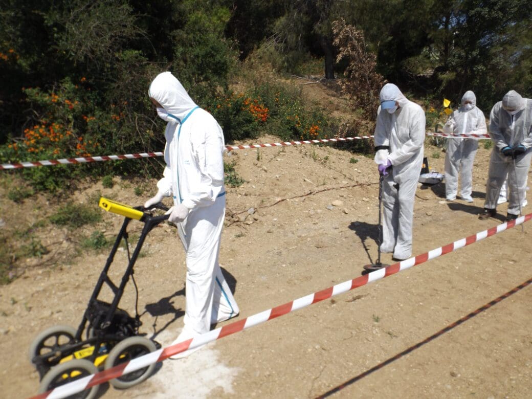 Forensic investigators in protective suits work behind police tape at an outdoor search scene, with evidence markers placed across the ground.