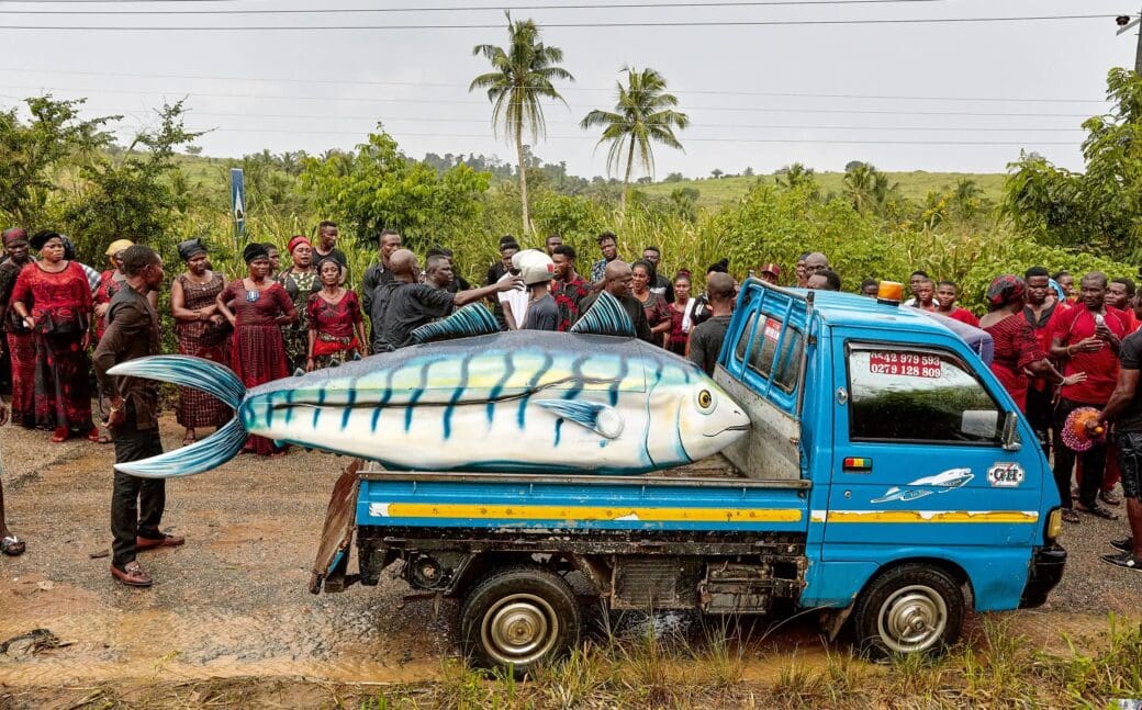 A fish-shaped fantasy coffin rests on the back of a blue truck, surrounded by mourners at a funeral in Ghana.