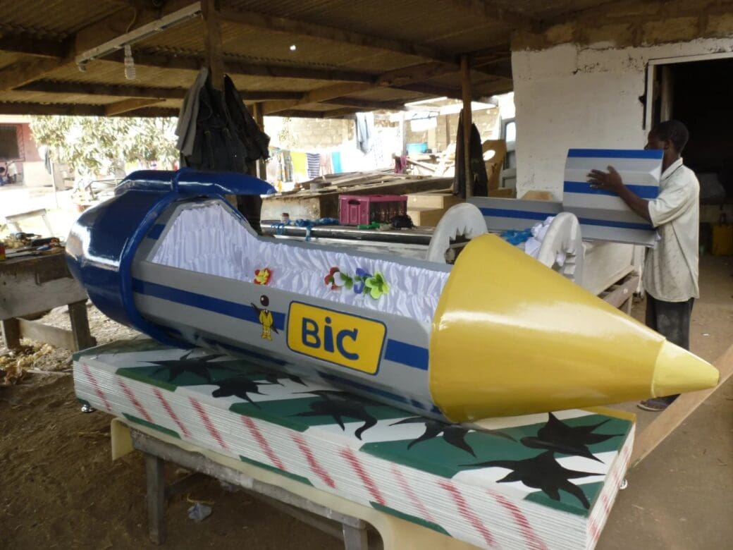 A large BIC pen-shaped fantasy coffin with a yellow tip and blue cap sits open inside a coffin workshop in Ghana.