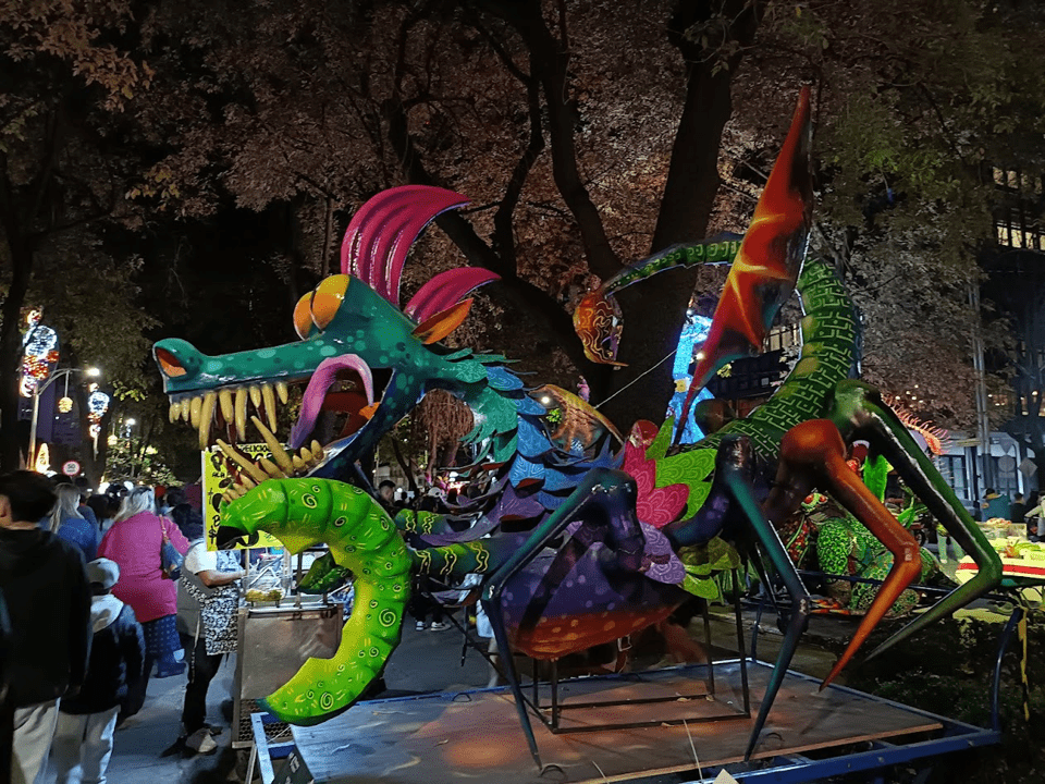  An alebrije statue displayed at night. This one looks to have a dragon's head which is green, with big round, orange eyes, a blue-purple-pink-green scaled body and a green, coiling tail.