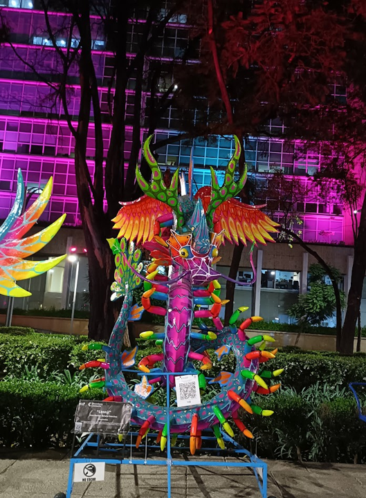 An alebrije statue displayed at night. This one has large orange wings, a purple face, round yellow eyes, multi-coloured legs and a blue tail.