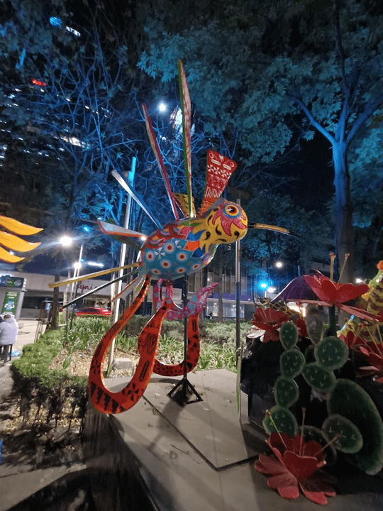 An alebrije statue displayed at night. It is yellow, pink and blue. It has hooked, orange legs and many colourful wings. It has round, wide eyes.