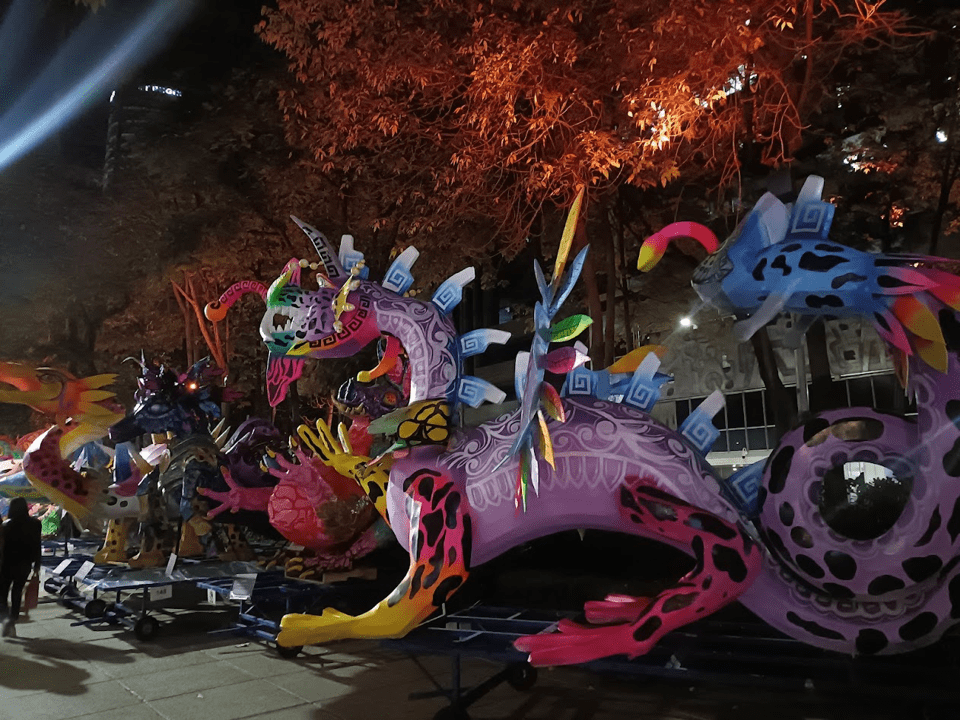 Several alebrije statues displayed at night. One has a dragon-like head, a large tail, square scales protruding from its back and spotted legs. Another is yellow and looks like a bird, with a long beak and yellow head feathers.