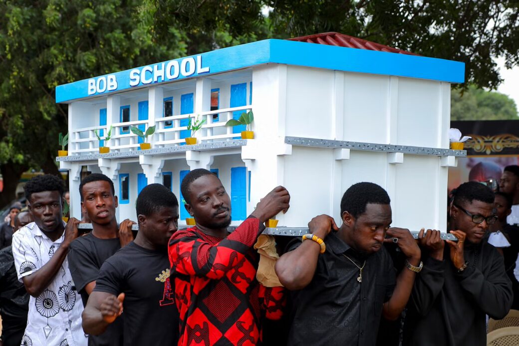 Pallbearers carry a fantasy coffin shaped like a white and blue school building with “BOB SCHOOL” written on the front.