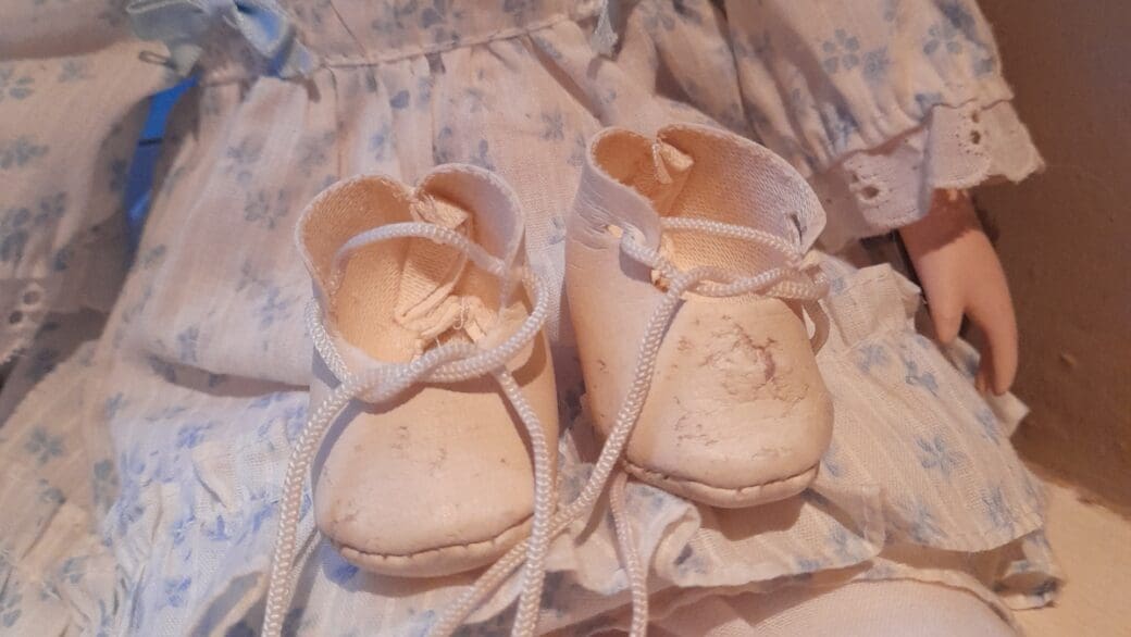 A pair of white, damaged doll shoes sit in the lap of a porcelain doll. The laces are undone. She's wearing a white dress wits arh faint blue flowers, and white lace ruffles on the sleeves.