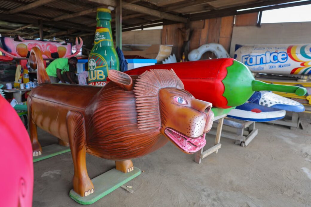 A lion-shaped fantasy coffin displayed alongside brightly painted coffins shaped like a pepper and other everyday objects.