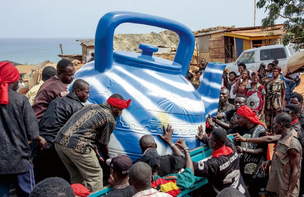 mourners gather around a large blue teapot-shaped fantasy coffin at a funeral in Ghana.