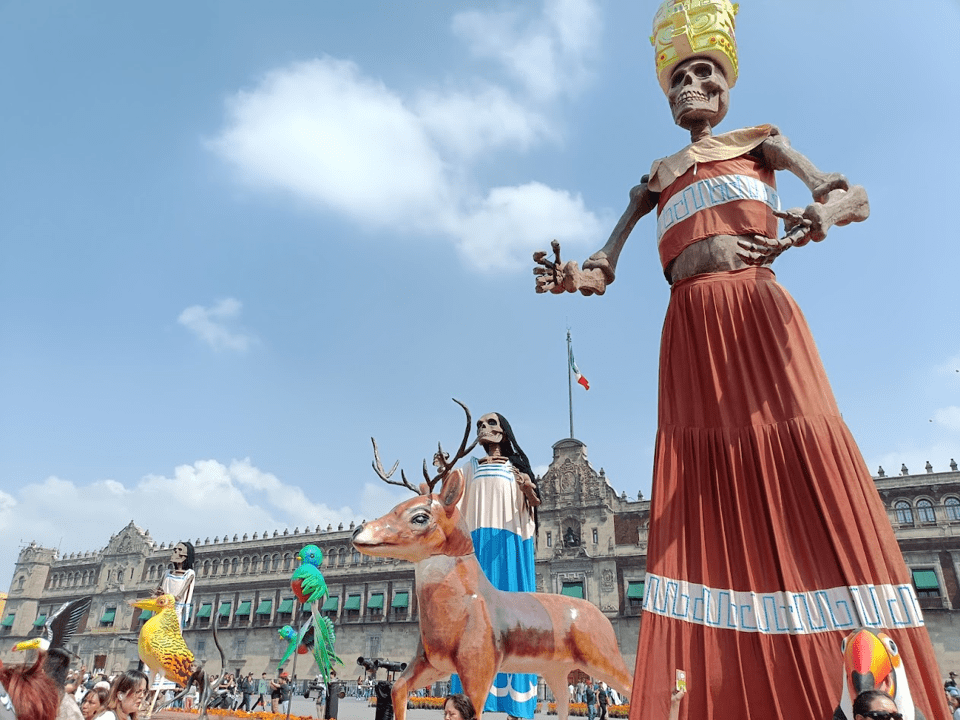 Celebrations in Zocalo Square, Mexico City. The sky is blue, with some clouds. There are large statues of skeletons outside a building, all of which are wearing long dresses or skirts.