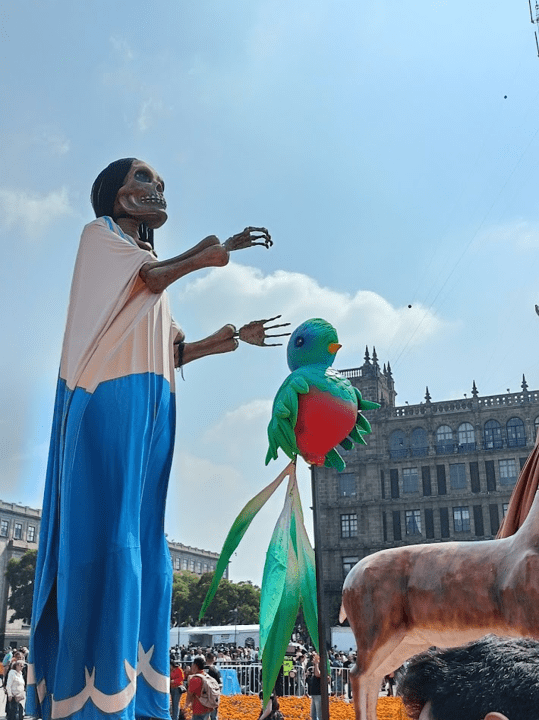 Celebrations in Zocalo Square, Mexico City. The sky is blue, with some clouds. There is a large statue of a skeleton, and another statue of a green and orange bird.