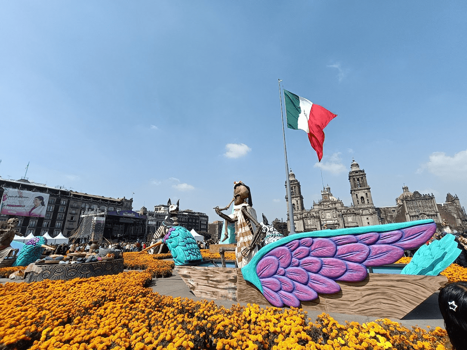 Celebrations in Zocalo Square, Mexico City. The sky is blue, with some clouds. There are orange flowers across the ground, and a boat with wooden purple-blue wings. Upon the boat is a skeleton and a Mexican flag. There is also a large statue of a dog.