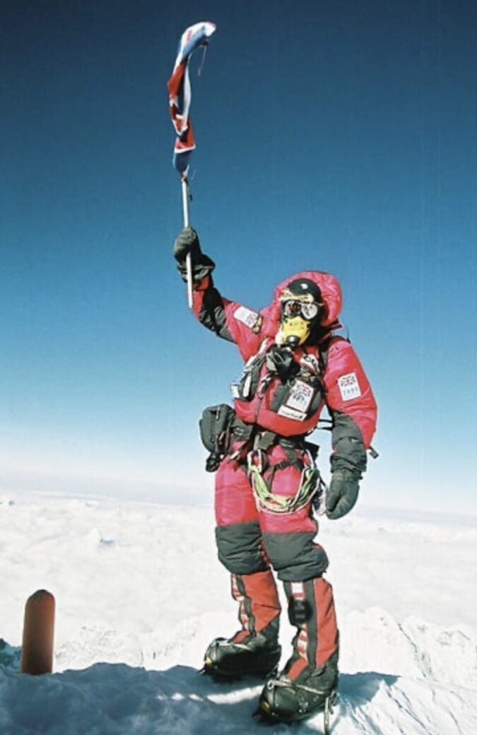 A climber in red summit gear raises a flag at the top of Everest against a clear blue sky.