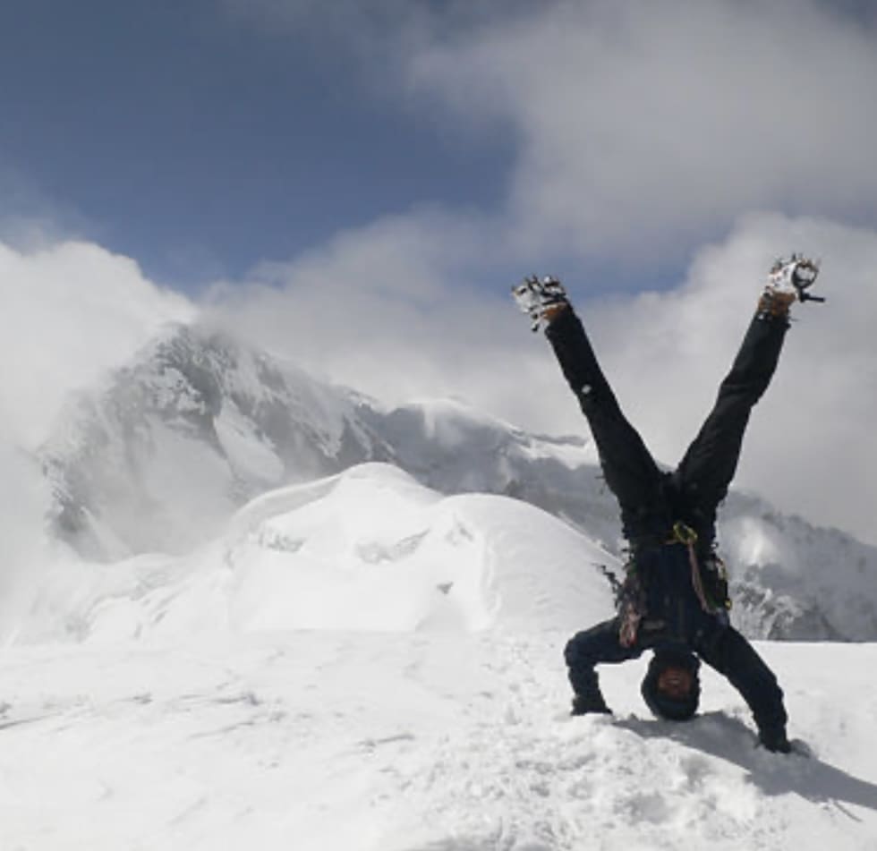 A climber performs a handstand on a snowy ridge with Everest partly hidden by cloud behind them.