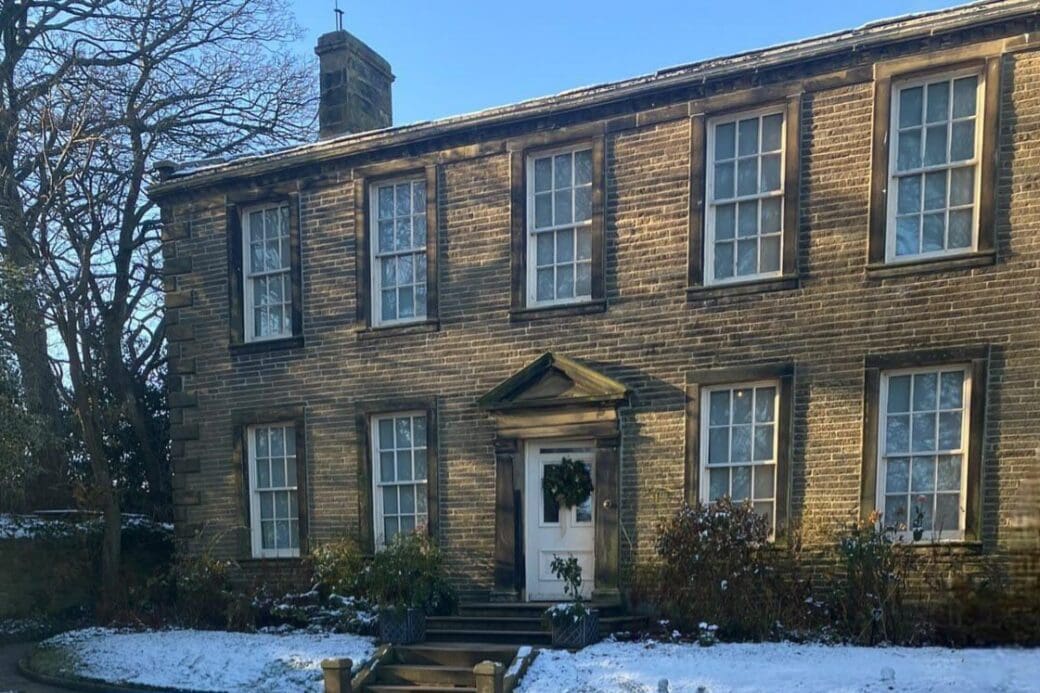 A brick building with several windows. It's winter and the trees are barren and snow is on the ground. There are steps leading up to a white door, which has a wreath on it.