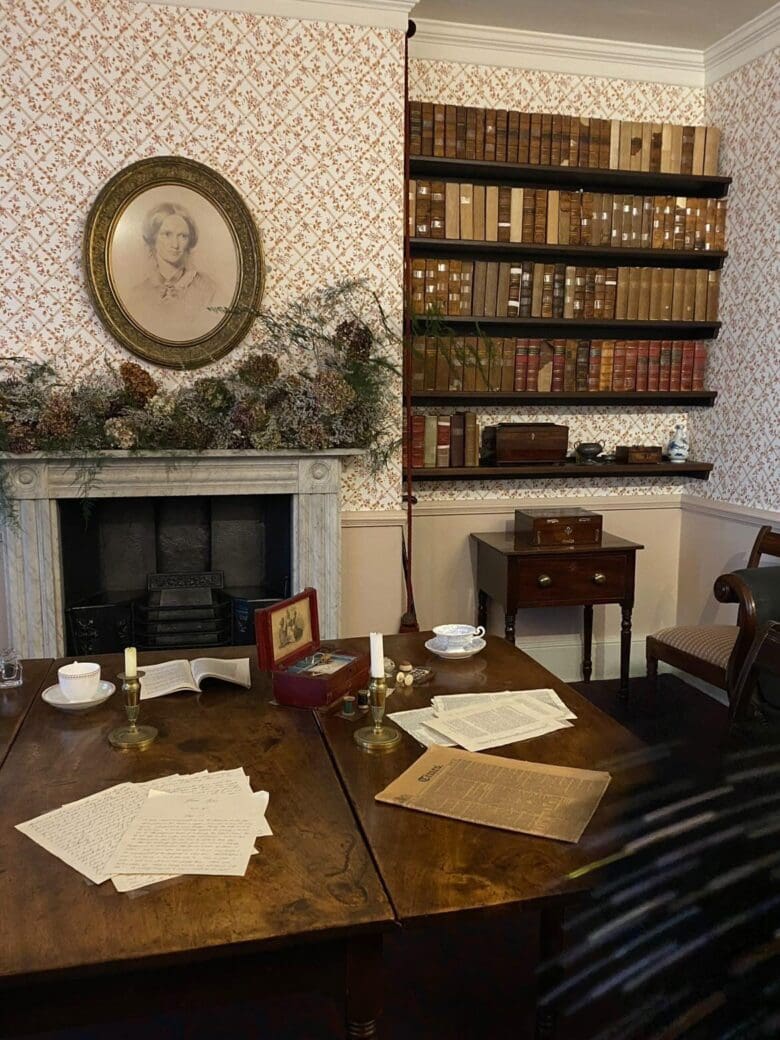 A dining room. It has floral wallpaper, a filled bookshelf, and a dark-wood table which has papers and candles on it. There is a stone fireplace and above it is a portrait.