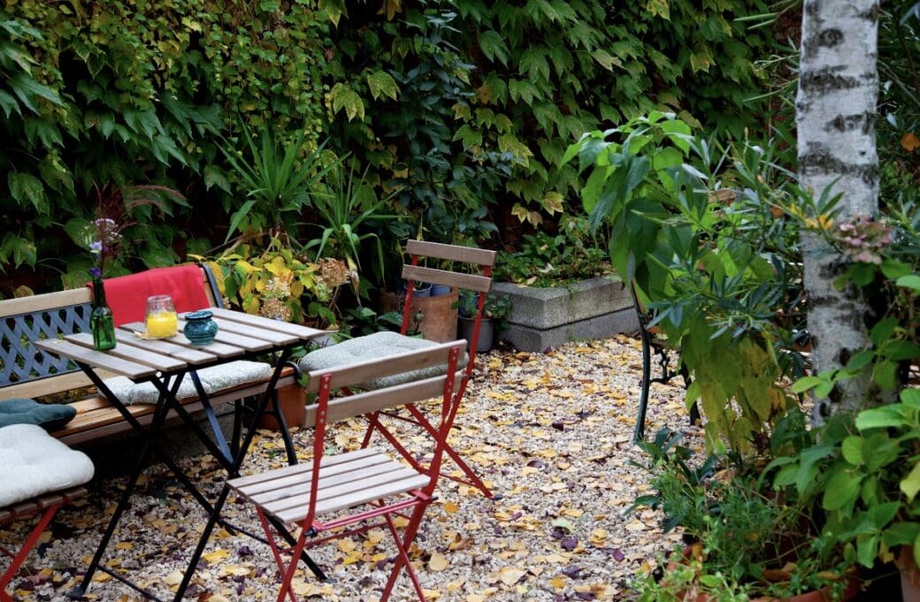 A sunny outdoor café garden with wooden tables and chairs set among trees, potted plants and gravel, beside a brick wall and pale building