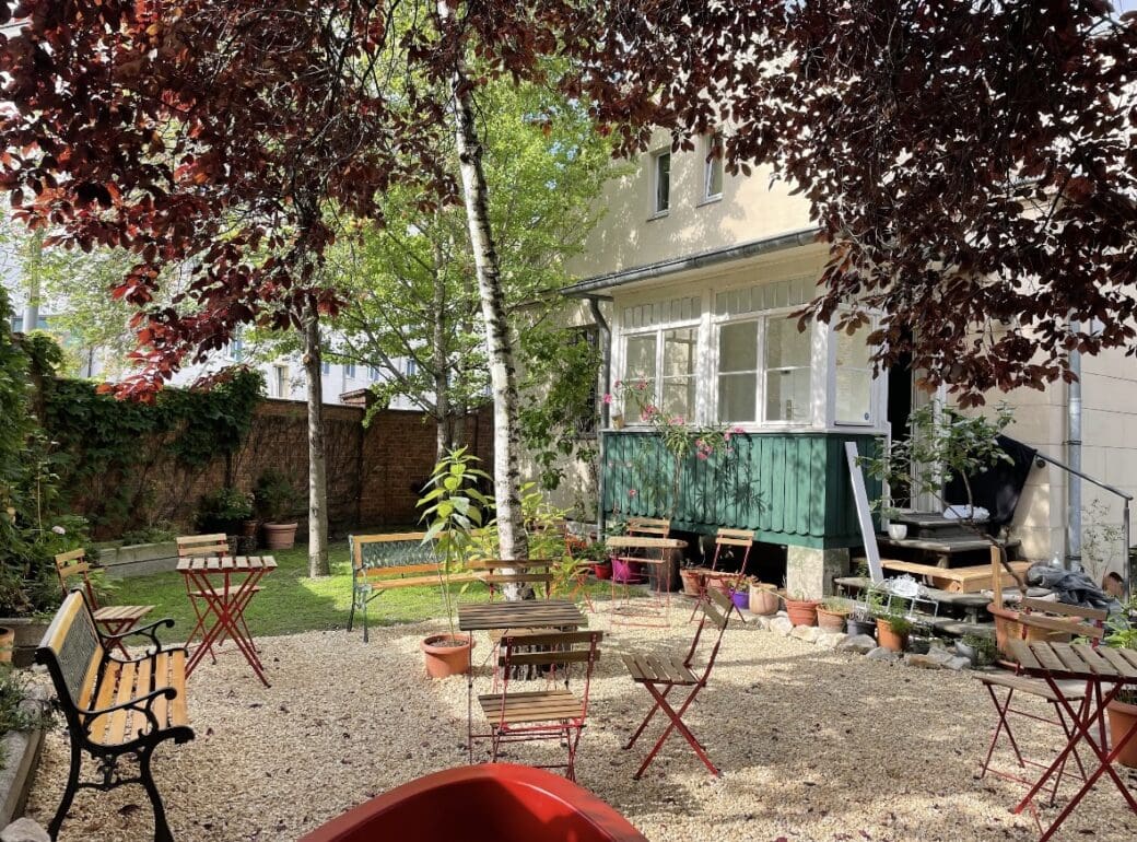 A sunny outdoor café garden with wooden tables and chairs set among trees, potted plants and gravel, beside a brick wall and pale building