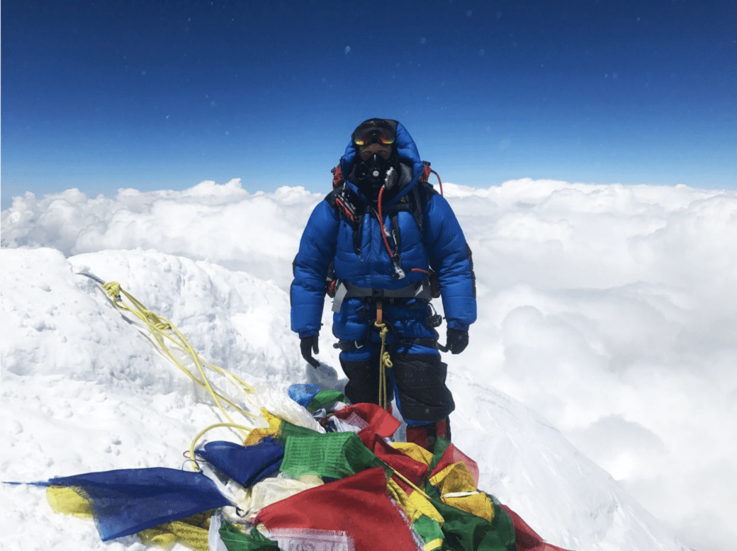 A climber in blue high-altitude gear stands on a snowy Himalayan ridge with Everest visible in the distance.