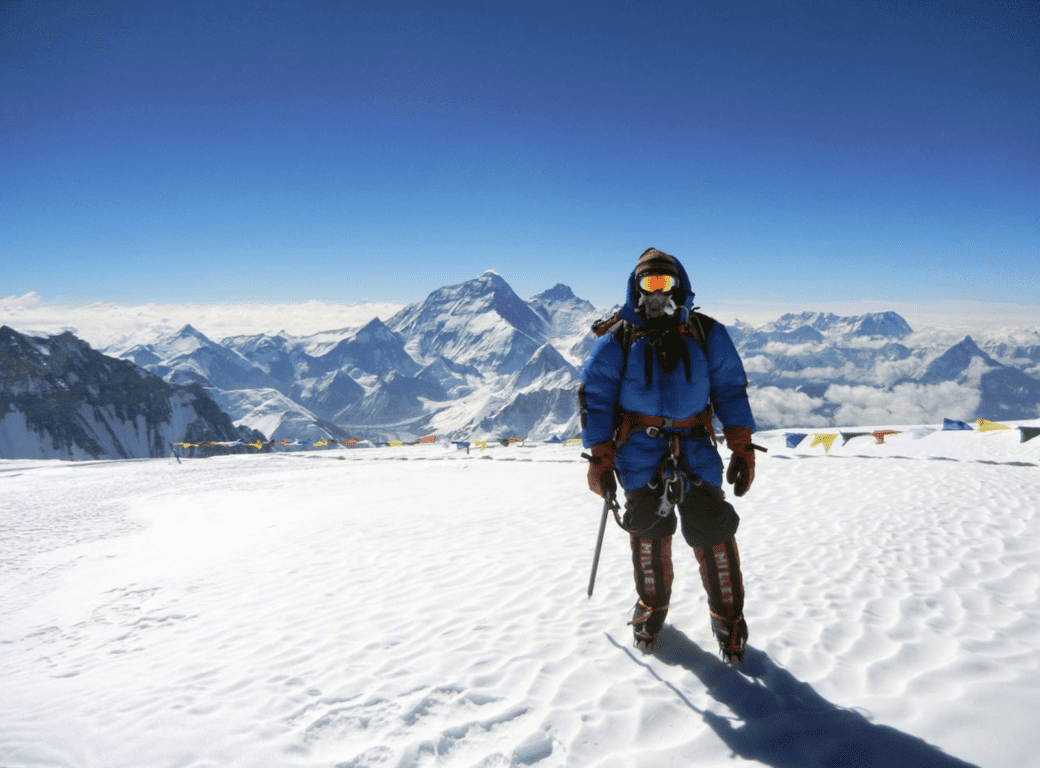 A climber in blue high-altitude gear stands on a snowy Himalayan ridge with Everest visible in the distance.