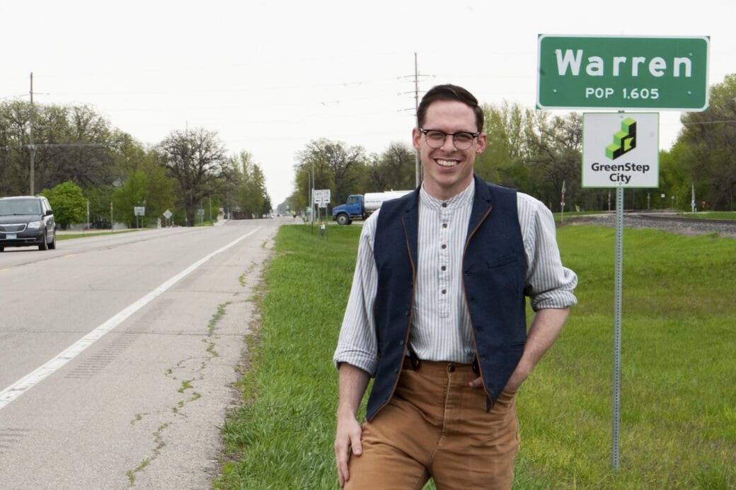 The man, Victor, standing in front of a sign which reads 'Warren, POP 1,605.' and 'GreenStop City.' He is wearing black glasses, a white shirt, a dark vest and brown pants. He is on the grass next to a road, where one black car passes by. The sky is bright and cloudy.