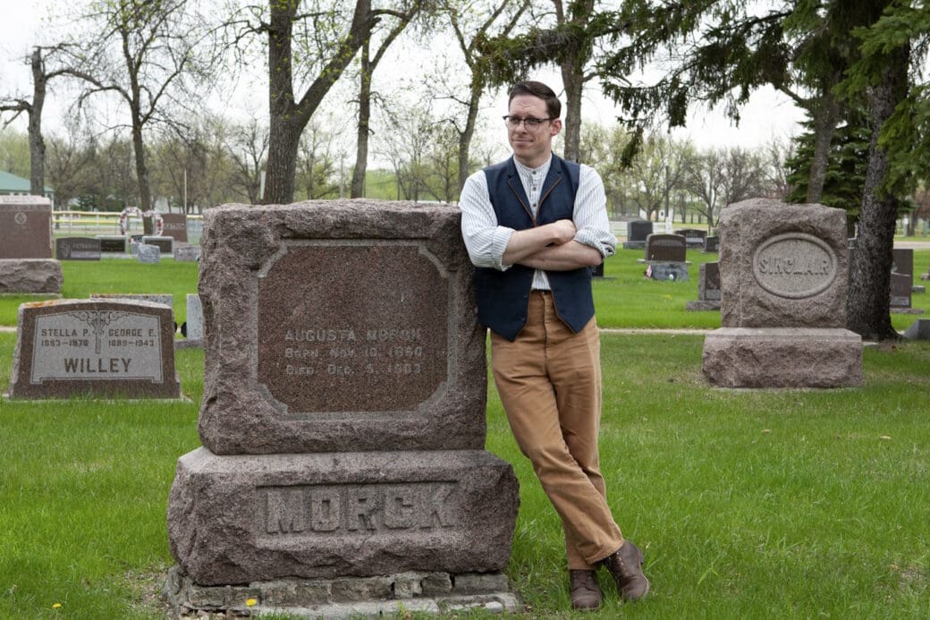 A man with glasses, a white shirt, a vest and brown pants leaning against a grave that reads 'MORCK.'