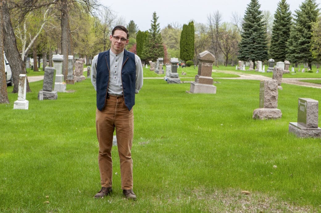 A man, Victor, wearing black glasses, a white shirt, a dark blue vest and brown pants. He is on the grass within a gravesite in front of several graves and trees. The sky is cloudy and bright white.