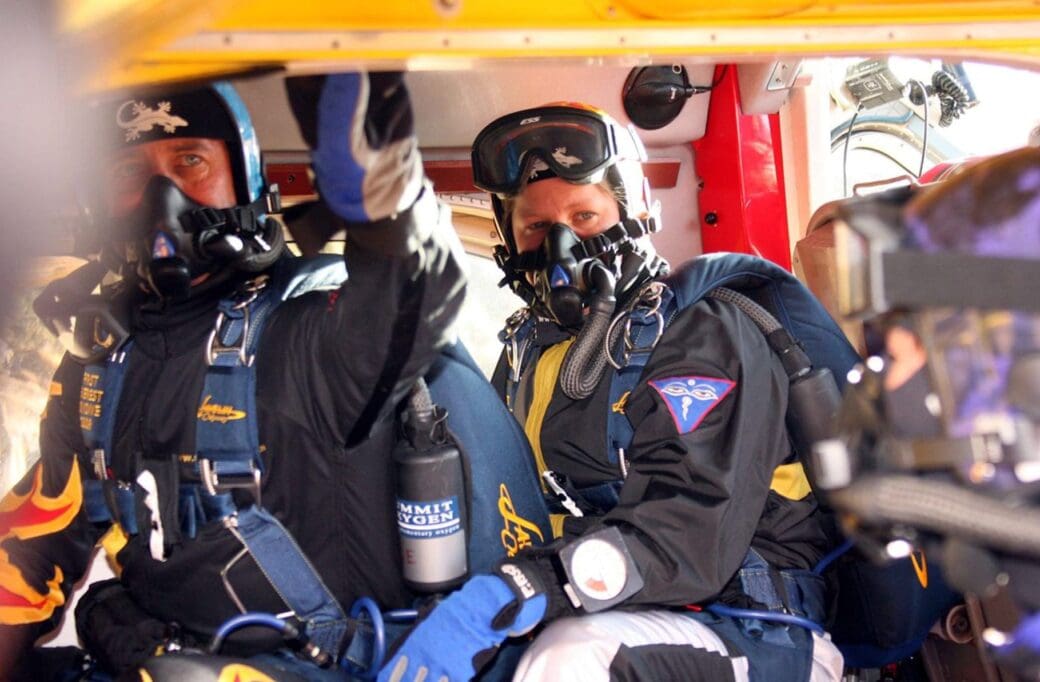 Two climbers wearing oxygen masks and skydiving gear sit inside an aircraft before a high-altitude jump near Everest.