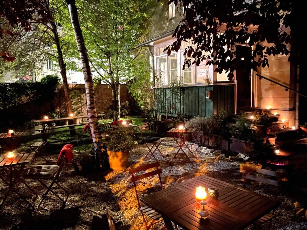 A candlelit outdoor café garden at night, with wooden tables and chairs set among trees, plants and gravel beside a warmly lit building.