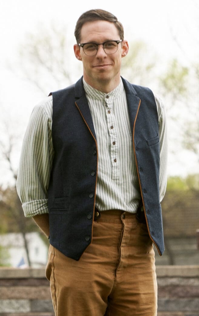 A white man wearing brown pants, a white shirt and a dark vest with glasses. He's standing in front of trees, and the sun is bright behind him. He's smiling at the camera.